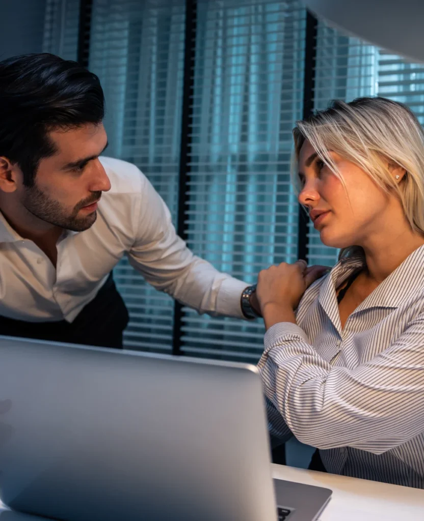 A man leans in towards a woman at a desk, both engaged in a serious discussion with a laptop nearby and blinds in the background.