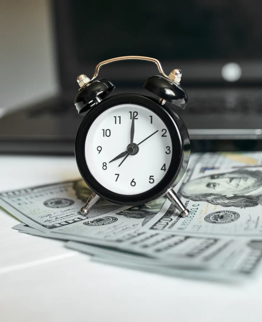 A black alarm clock on a stack of hundred-dollar bills, with a blurred laptop in the background, symbolizing time and money.