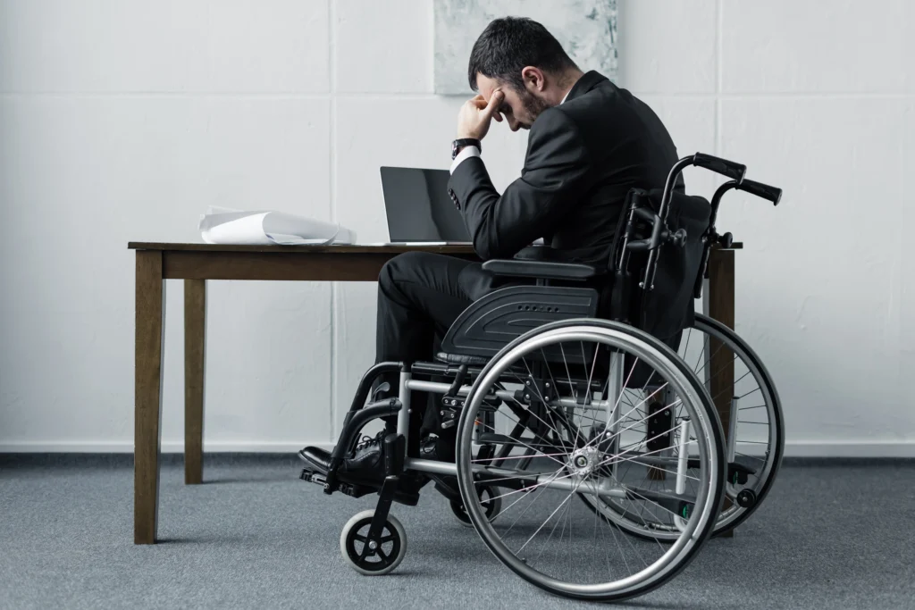 A businessman in a wheelchair sits at a desk, resting his head on his hand, with a laptop and papers nearby.