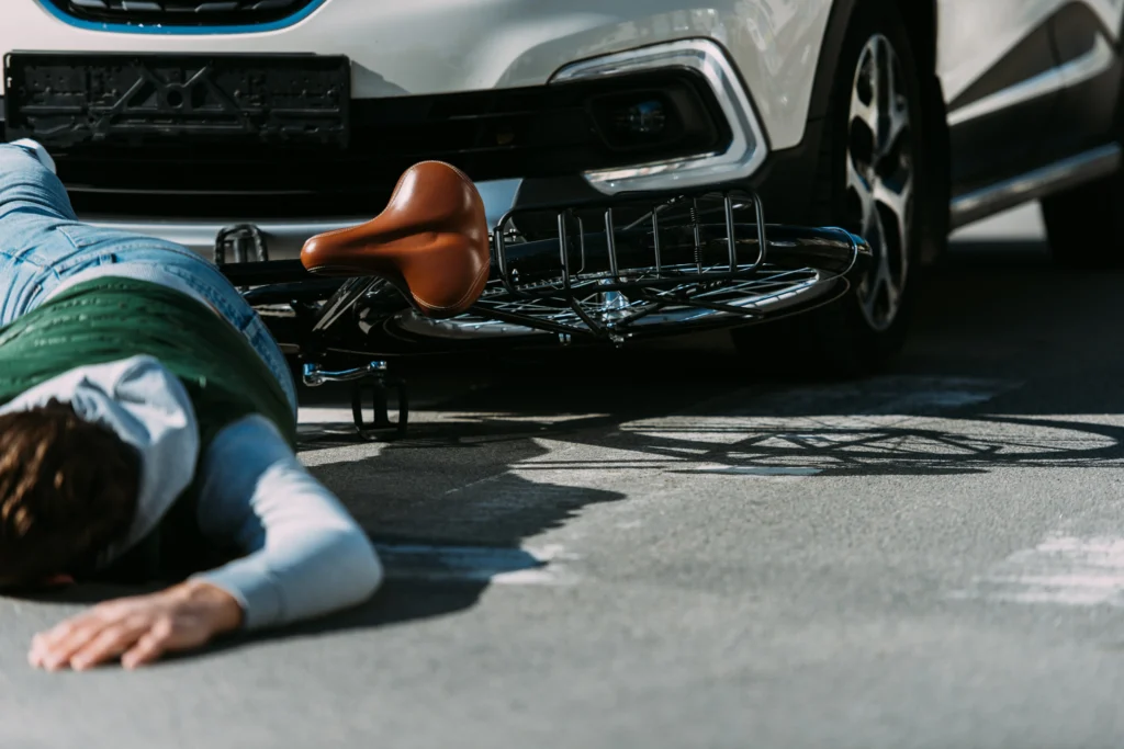 A cyclist lies injured on the ground near a white car, with a bicycle nearby, highlighting a serious accident scene.