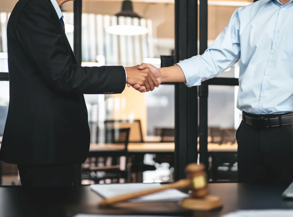 Two businessmen in formal attire shake hands in an office setting, with a gavel in the foreground, symbolizing agreement or partnership.