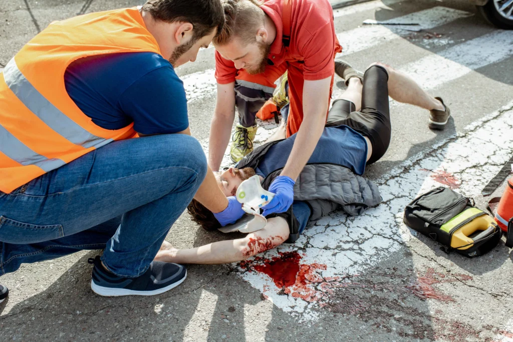 A small group of people sitting and painting on the sidewalk, surrounded by various art supplies. They are dressed casually in jeans and t-shirts, with a car parked nearby on the street.