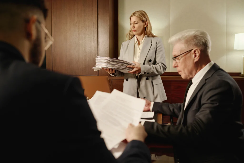 A woman in a gray suit holds a stack of documents in an office meeting with two men focused on paperwork in front of them.