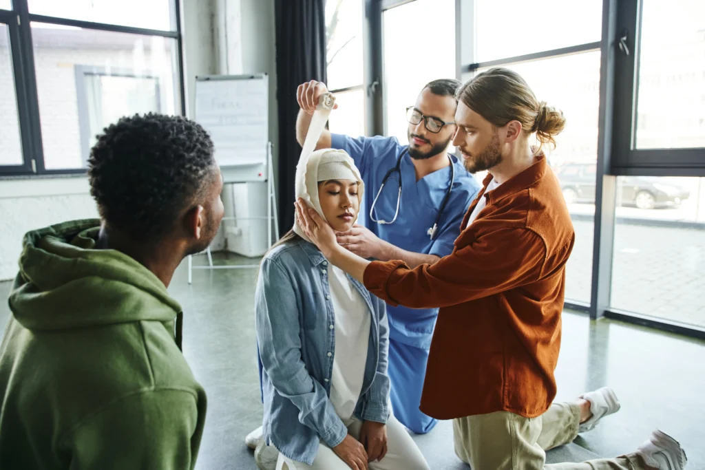 A medical training scenario where a person is demonstrating how to apply a bandage on another's head, with onlookers observing.