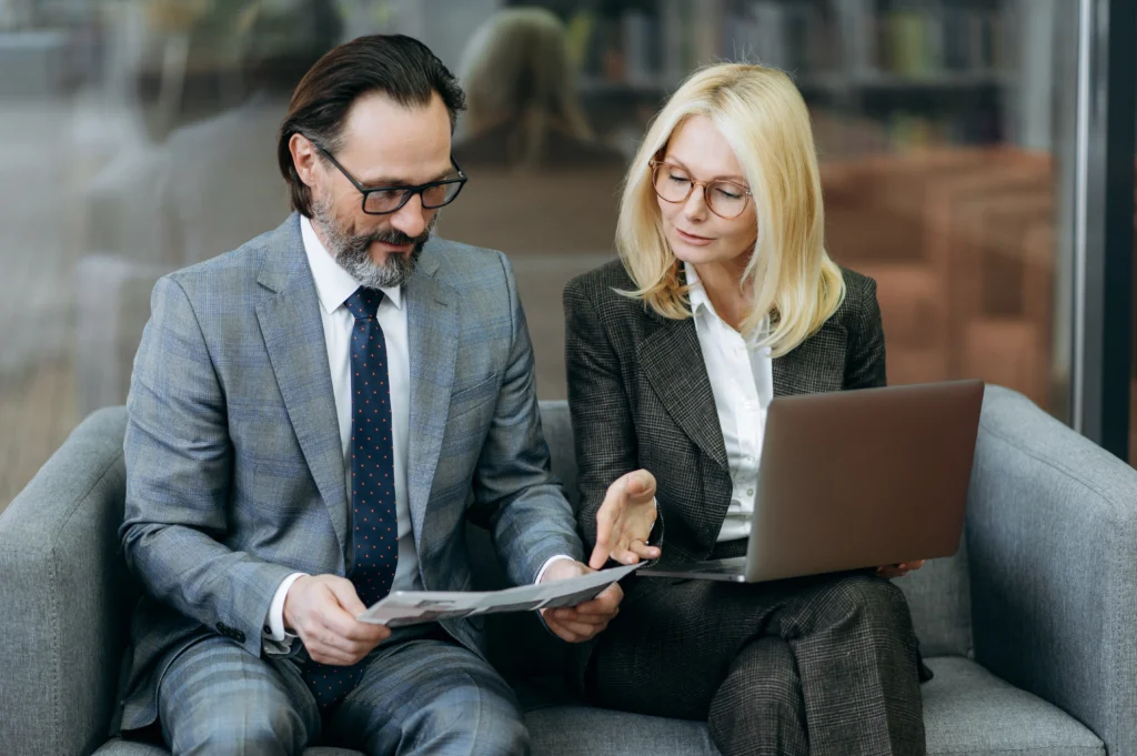 Two business professionals engaged in a discussion, one holding documents and the other using a laptop, seated on a gray couch.