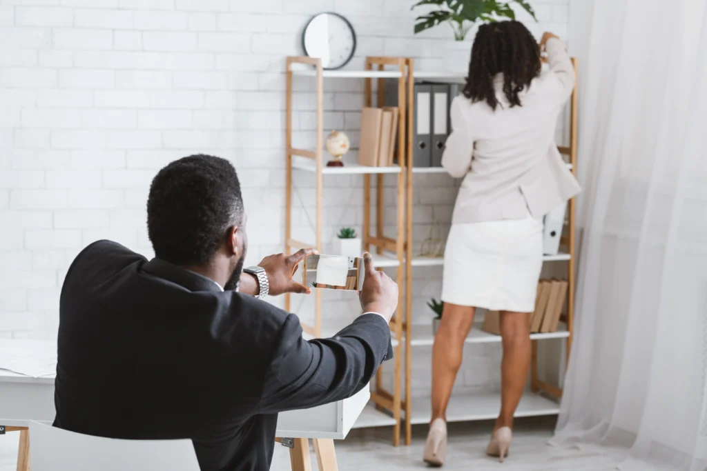A man in a suit captures a photo of a woman in a light blazer, as she organizes items on an office shelf.
