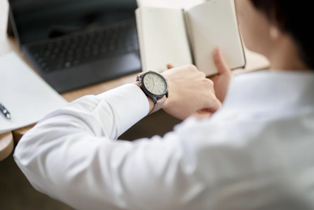 A person in a white shirt checks their watch while holding an open notebook, with a laptop and papers on the desk in the background.