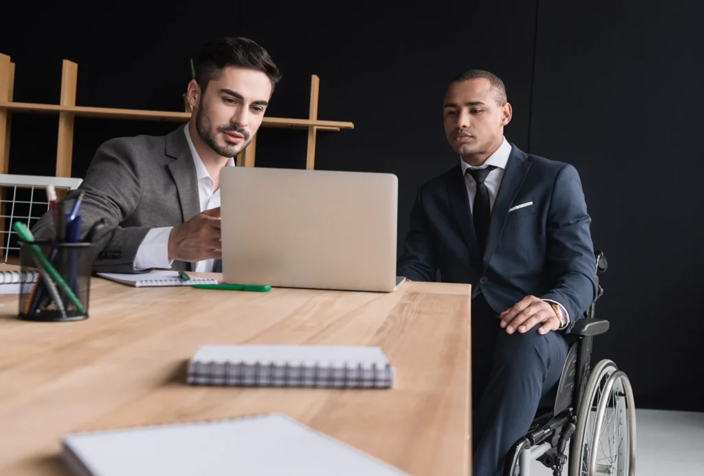 Two professionals engaged in discussion at a wooden table, with a laptop and notebooks in a modern office setting.