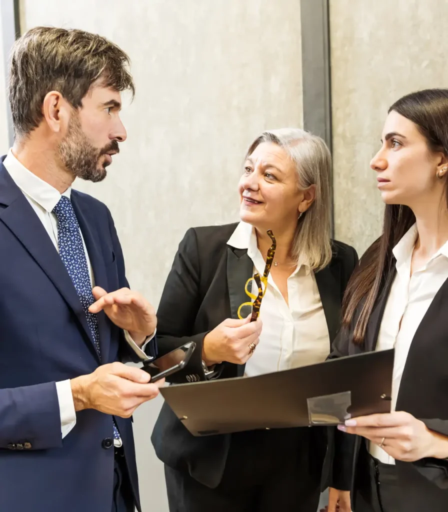 Three professionals in business attire engage in a discussion. A man gestures with a phone, while two women hold documents, appearing focused and collaborative.