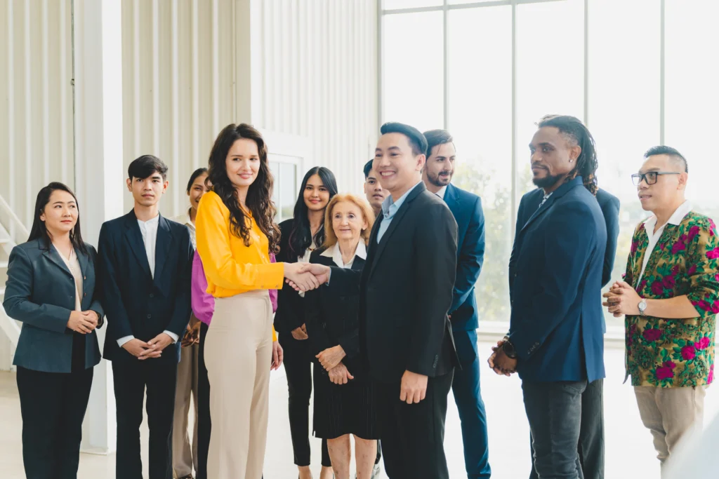 A diverse group of professionals in formal attire stands in a bright office. Two individuals in the center shake hands, smiling, conveying a sense of agreement and teamwork.