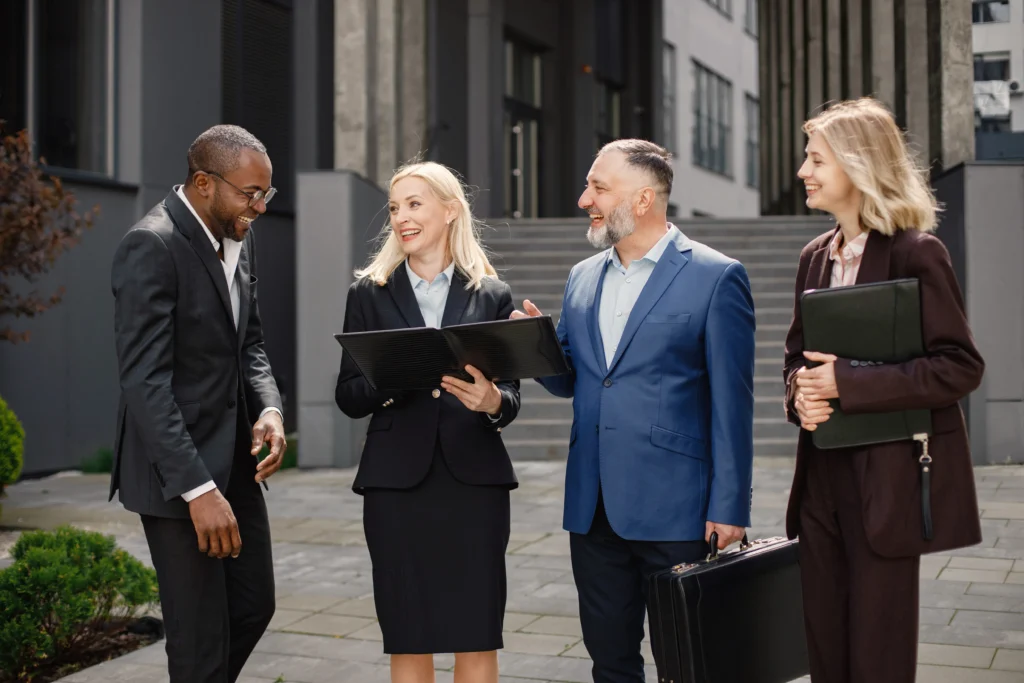Four business professionals in formal attire stand outside, engaged in conversation. They appear cheerful and collaborative, holding briefcases and documents.