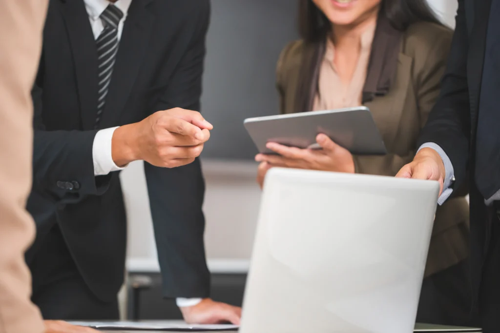 Business meeting with three professionals in formal attire. One is gesturing at a laptop screen while another holds a tablet, conveying collaboration.