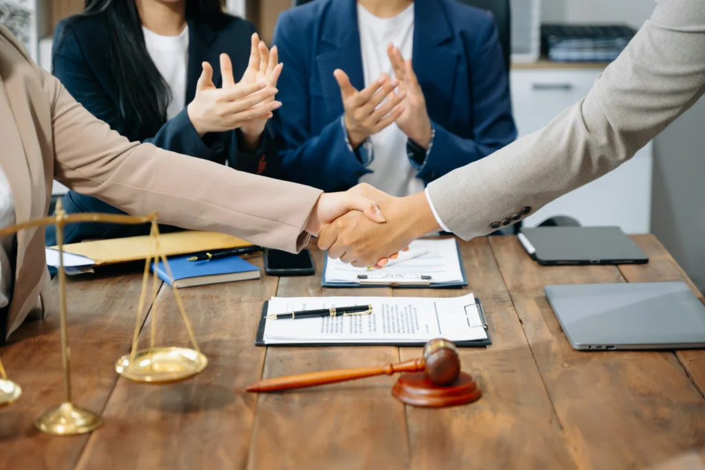 Two people in business attire are shaking hands over a table with a gavel and scales, signifying an agreement. Colleagues in the background are applauding.