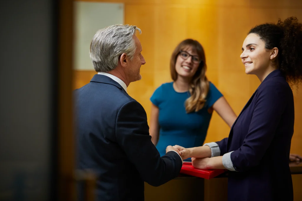 A woman in a blue dress smiles as two professionals in suits shake hands, conveying a warm, welcoming business environment.