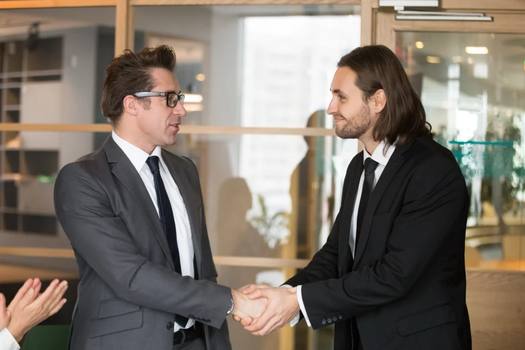 Two men in suits shake hands in a professional setting, conveying agreement and mutual respect. A woman on the left applauds, showing approval.