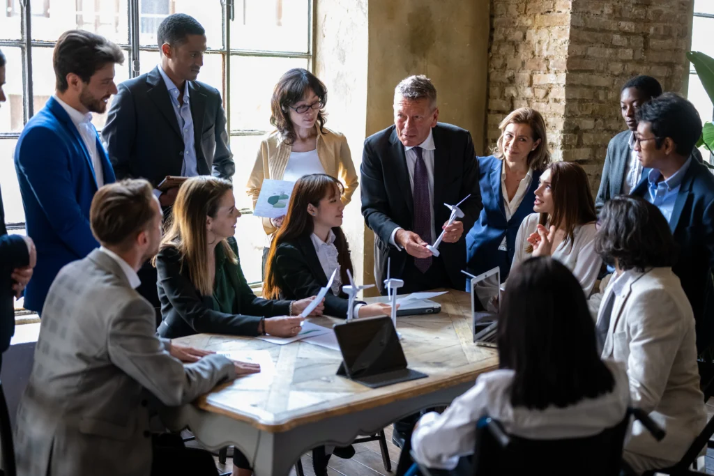 A diverse group of professionals in formal attire are engaged in a lively discussion around a table, holding papers and miniature wind turbines, suggesting a focus on renewable energy in a collaborative office setting.