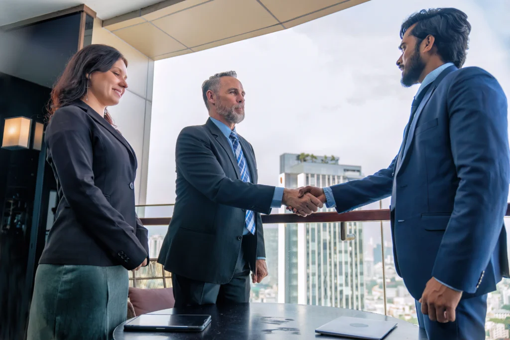 Two businessmen in suits shake hands confidently on a balcony with a cityscape backdrop. A woman in business attire stands beside them, smiling.