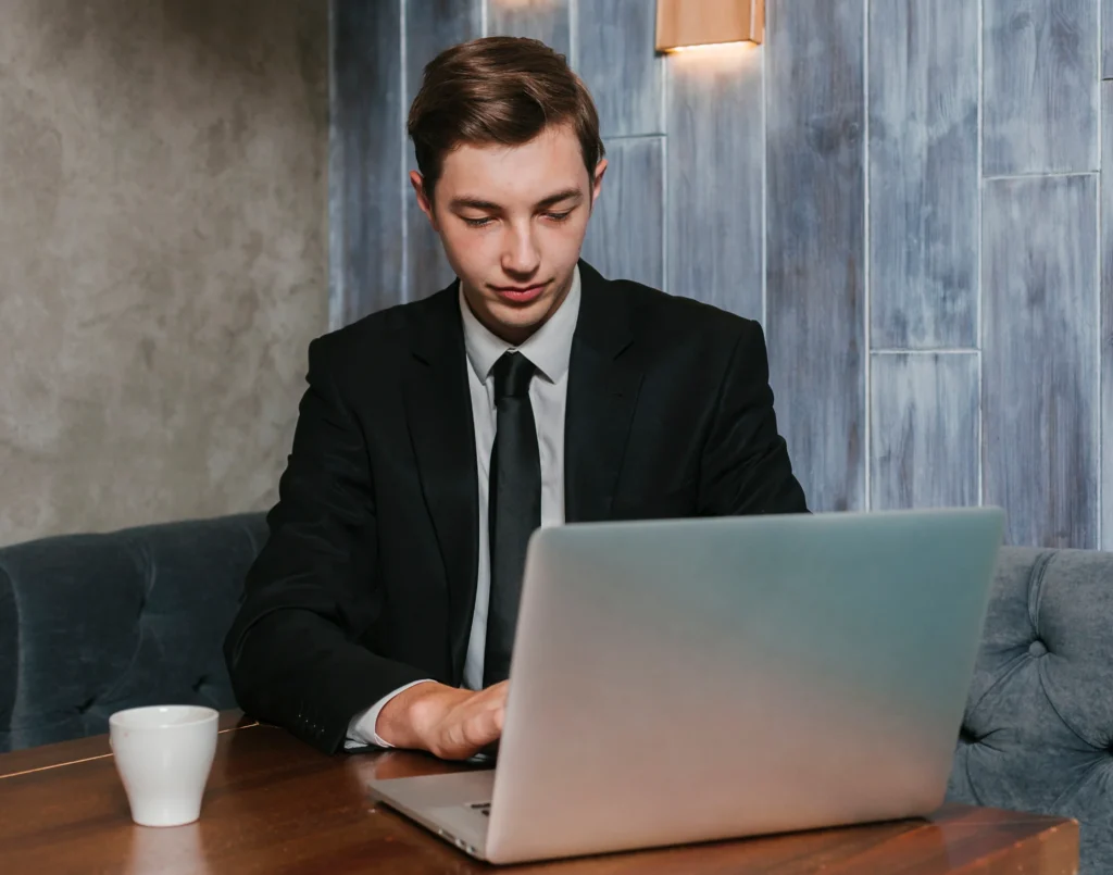 Young man in a suit works on a laptop at a wooden table in a cozy cafe. A white mug sits beside him. The atmosphere feels focused and professional.