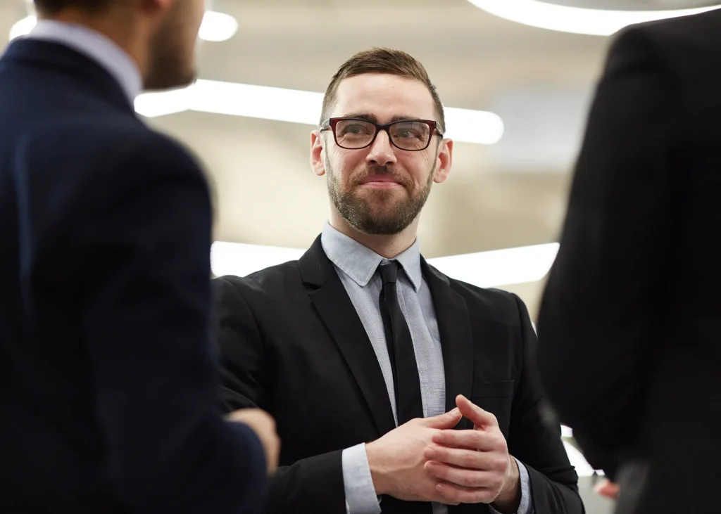 A man in a suit, wearing glasses, stands confidently in an office setting, listening attentively to colleagues. The atmosphere is professional and engaged.