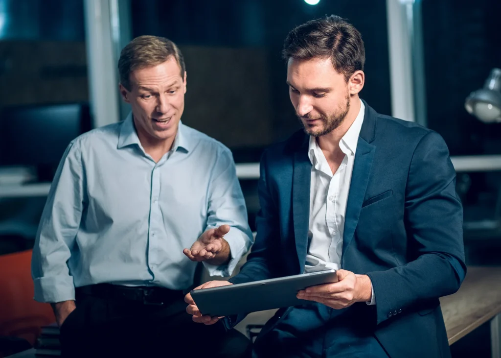Two men in an office setting: one in a light blue shirt gesturing while speaking, and the other in a dark suit attentively looking at a tablet.
