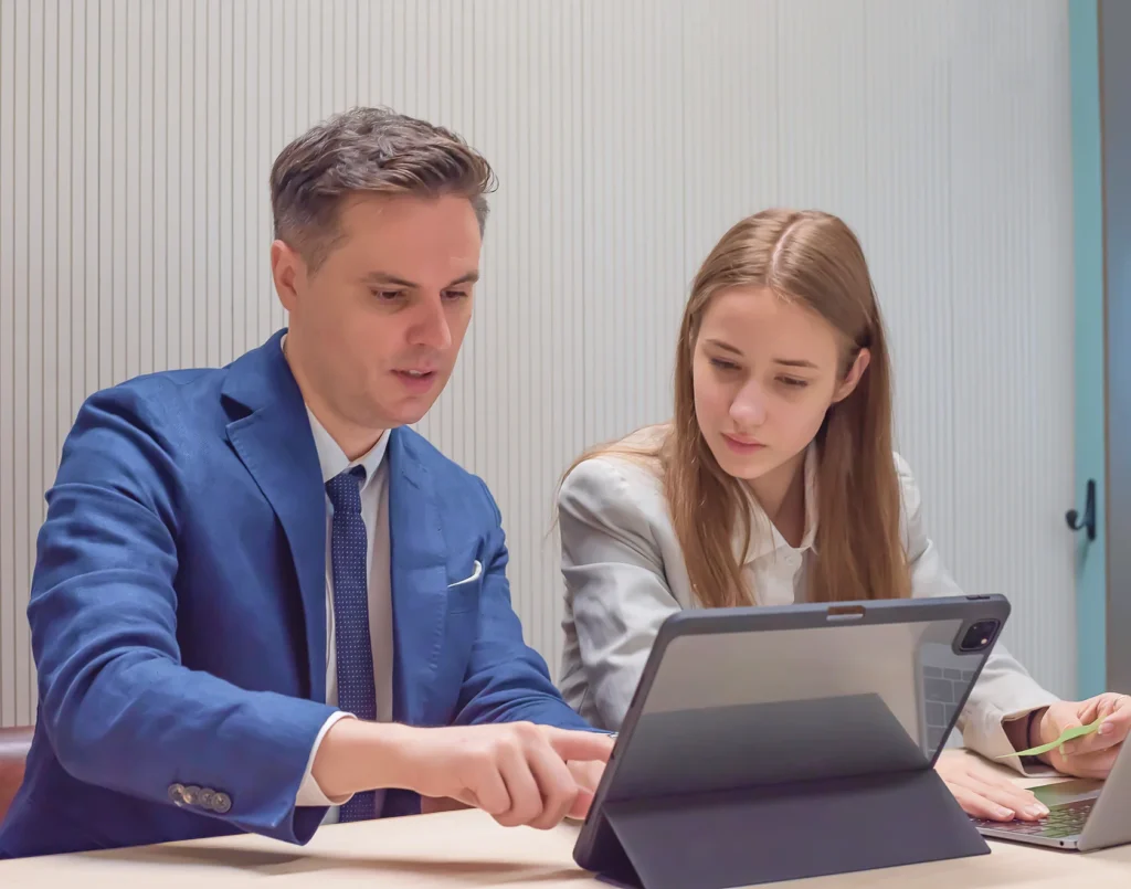 A man in a blue suit and a woman in a light blazer collaborate over a tablet during a business meeting.
