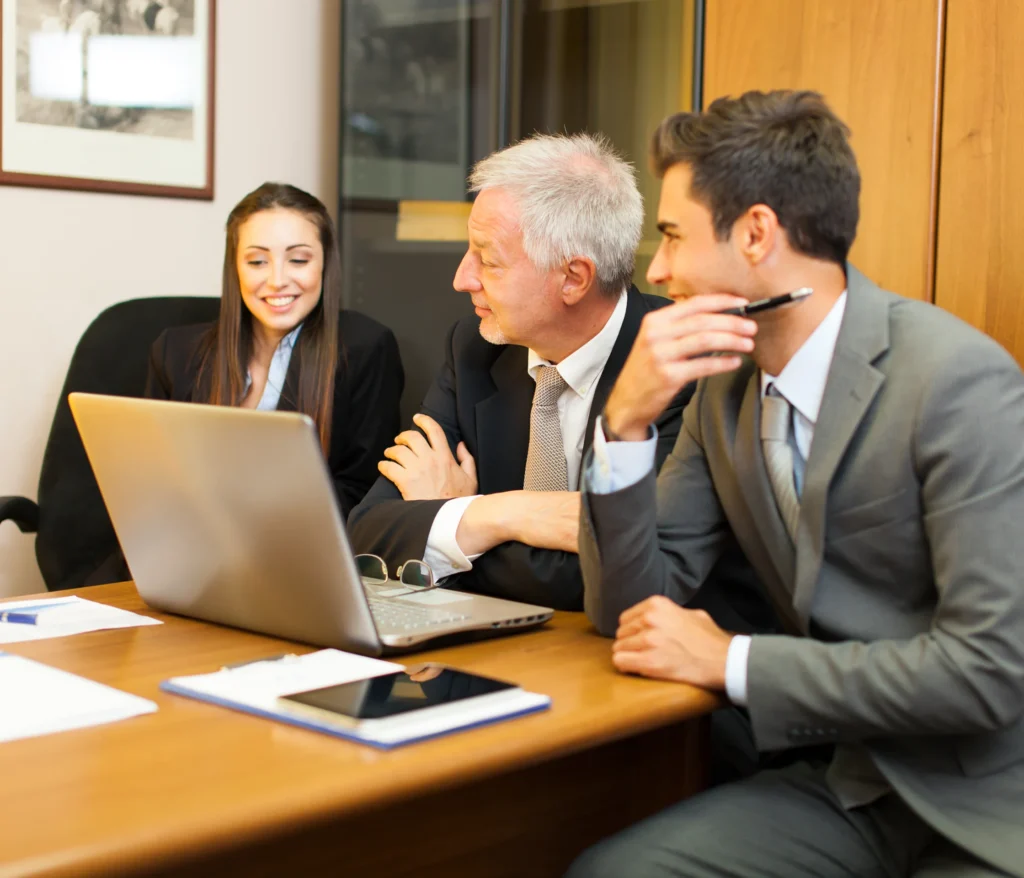 Three business professionals in formal attire, two men and one woman, engaged in a discussion around a laptop in a modern office setting. They appear focused and collaborative.