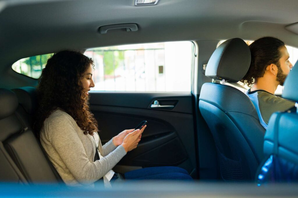 A woman sitting in the back seat of a rideshare vehicle, looking at her phone while the driver focuses on driving.
