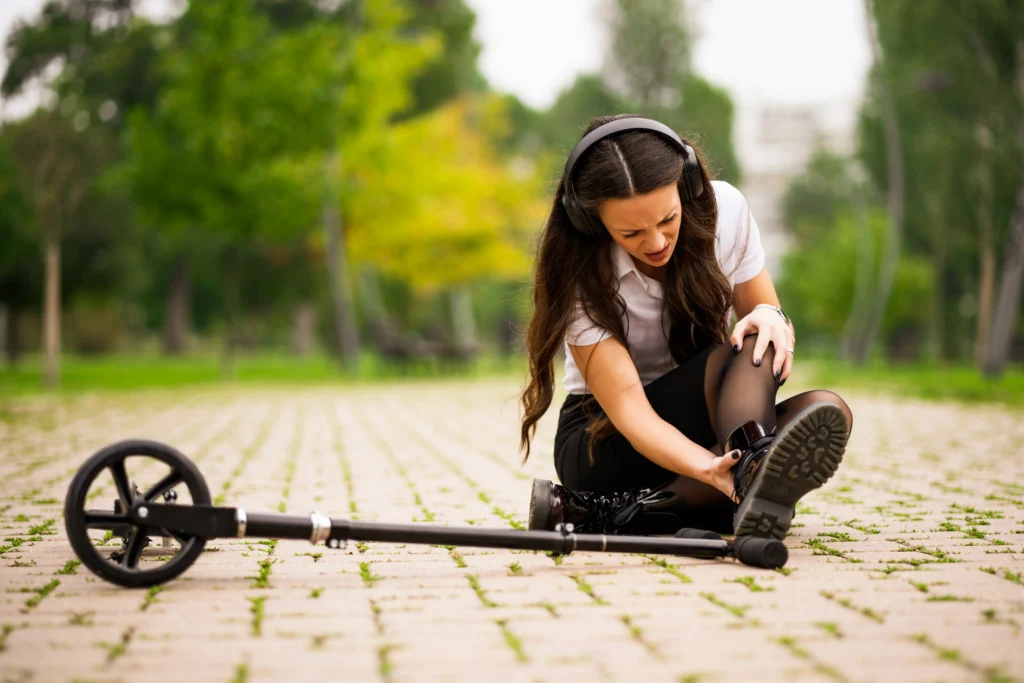 Young woman wearing headphones is seated outdoors, adjusting her shoe in a park, with greenery and trees in the background.