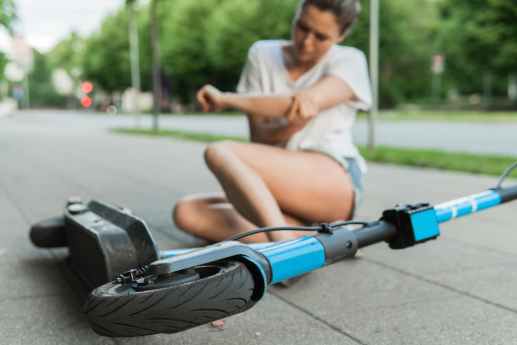 Woman sits on the ground, holding her arm in pain, near a fallen blue electric scooter on the sidewalk
