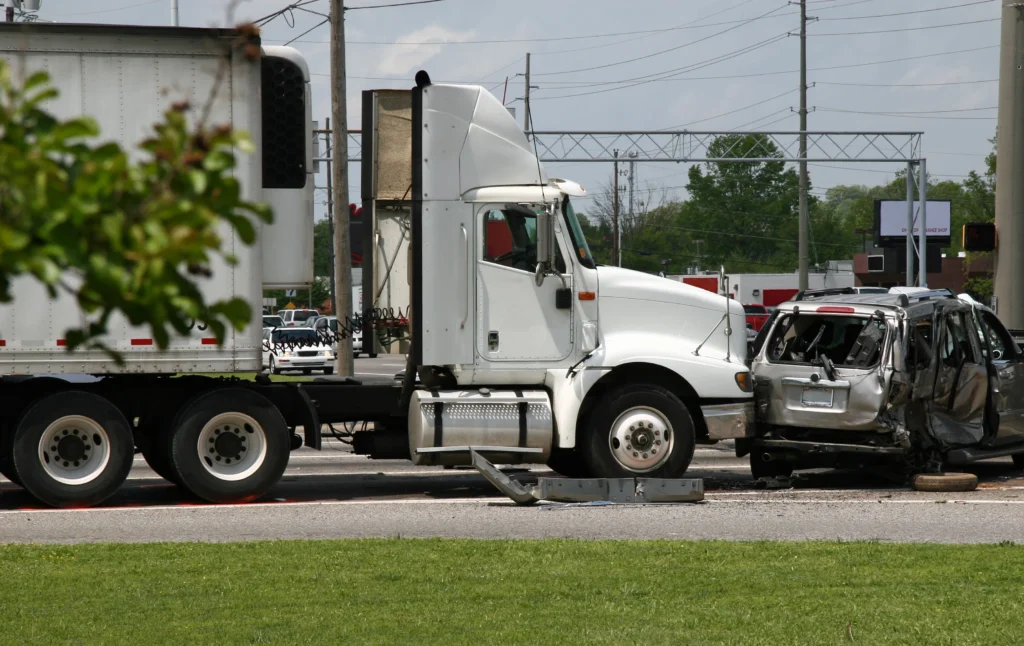 Damaged silver SUV is crushed against the front of a white semi-truck, with debris scattered on the road in a busy intersection.