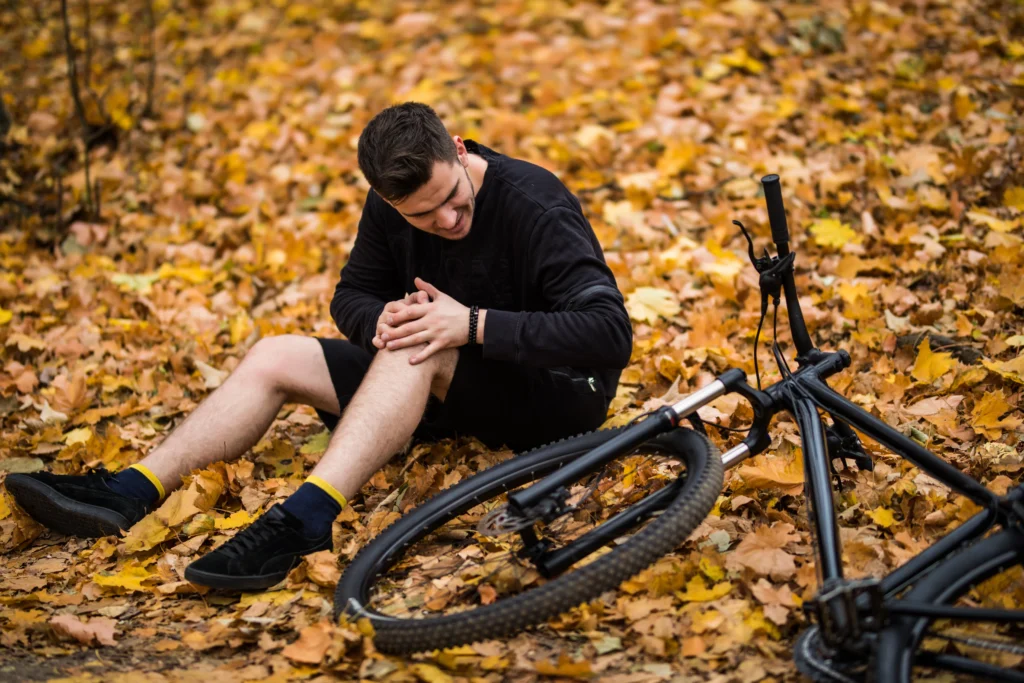 A person sits on a bed of autumn leaves, holding their knee, with a black bike lying on the ground nearby.