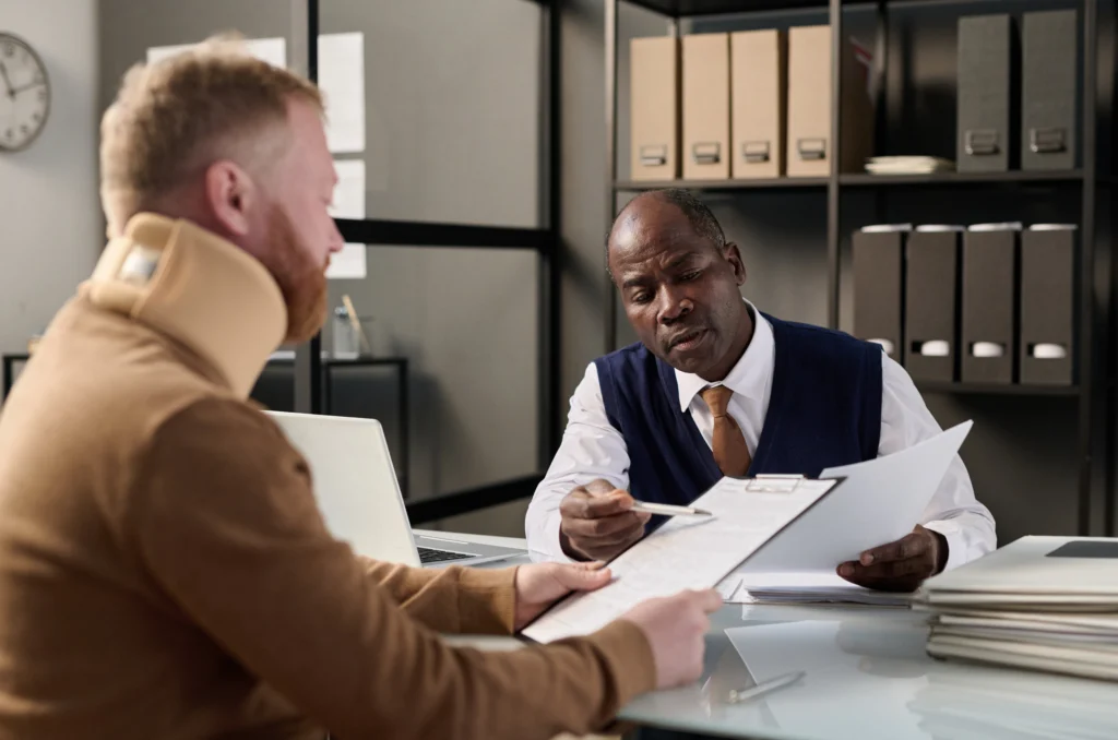 A professional meeting in an office setting, with two individuals discussing documents at a glass table surrounded by file cabinets.