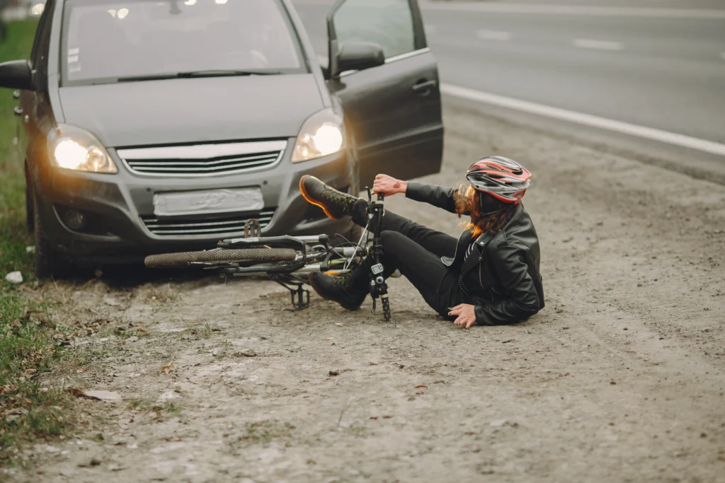 A person in cycling gear sits on the ground beside a fallen bicycle, with a car parked nearby and its headlights on.