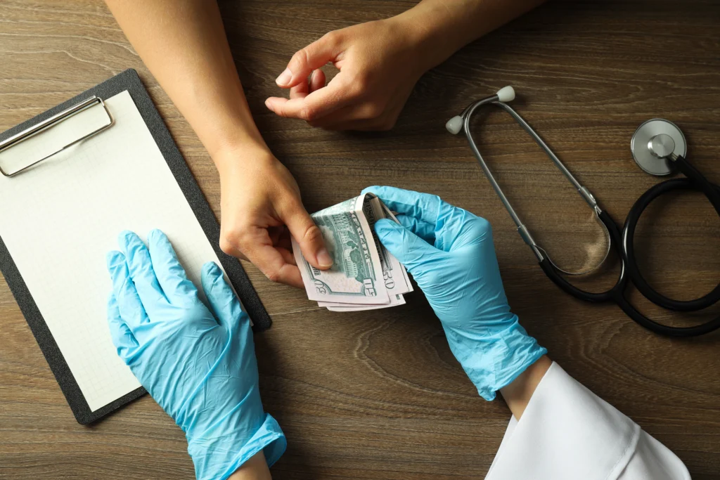 A gloved hand exchanges cash with another hand, beside a clipboard and stethoscope on a wooden table.