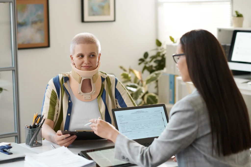 An individual wearing a neck brace sits at a desk, while another person gestures towards a laptop with documents. Plants and art are in the background.