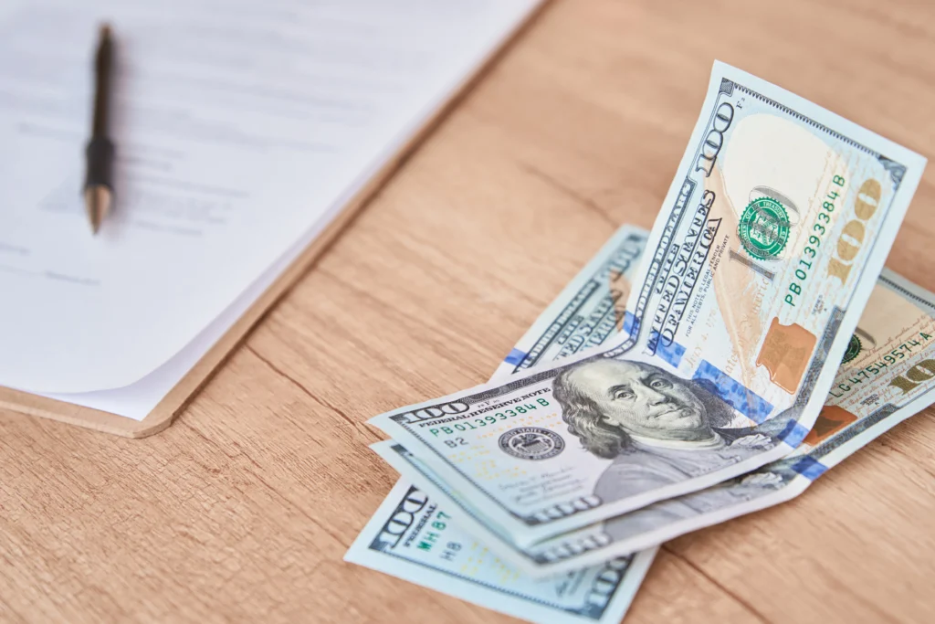 A pile of hundred-dollar bills on a wooden table next to a stack of documents and a black pen.