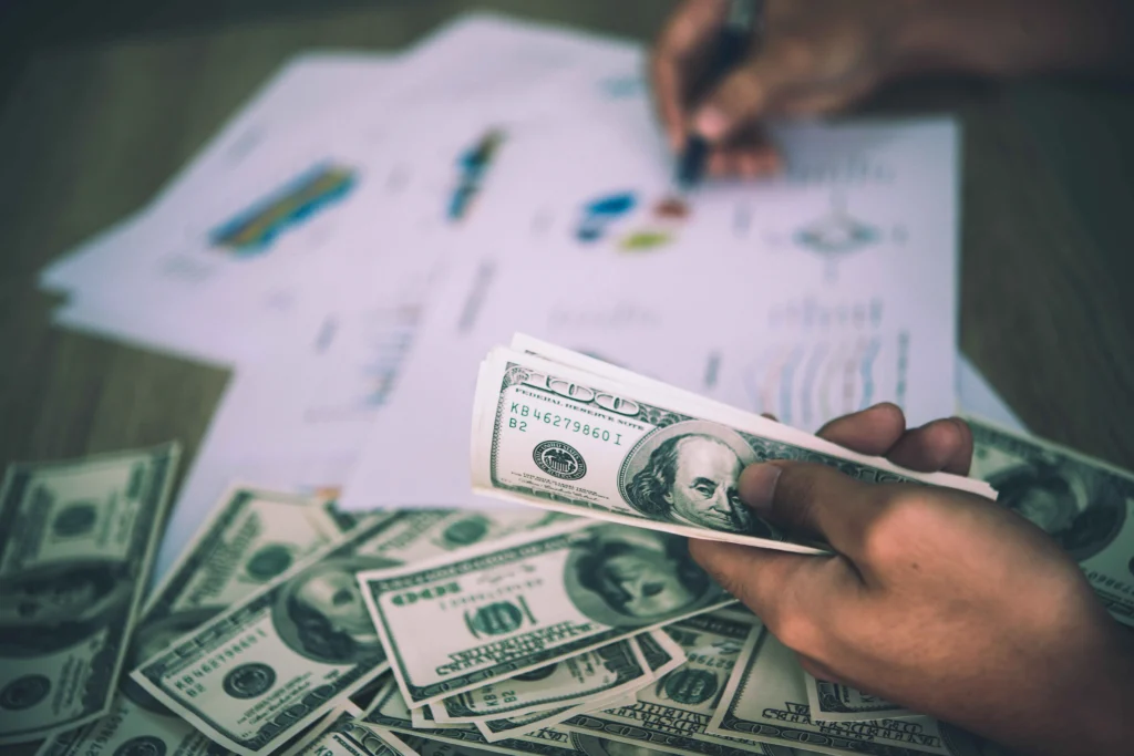 A hand holds a stack of bills over a scattered pile of cash, with financial documents and charts in the background.