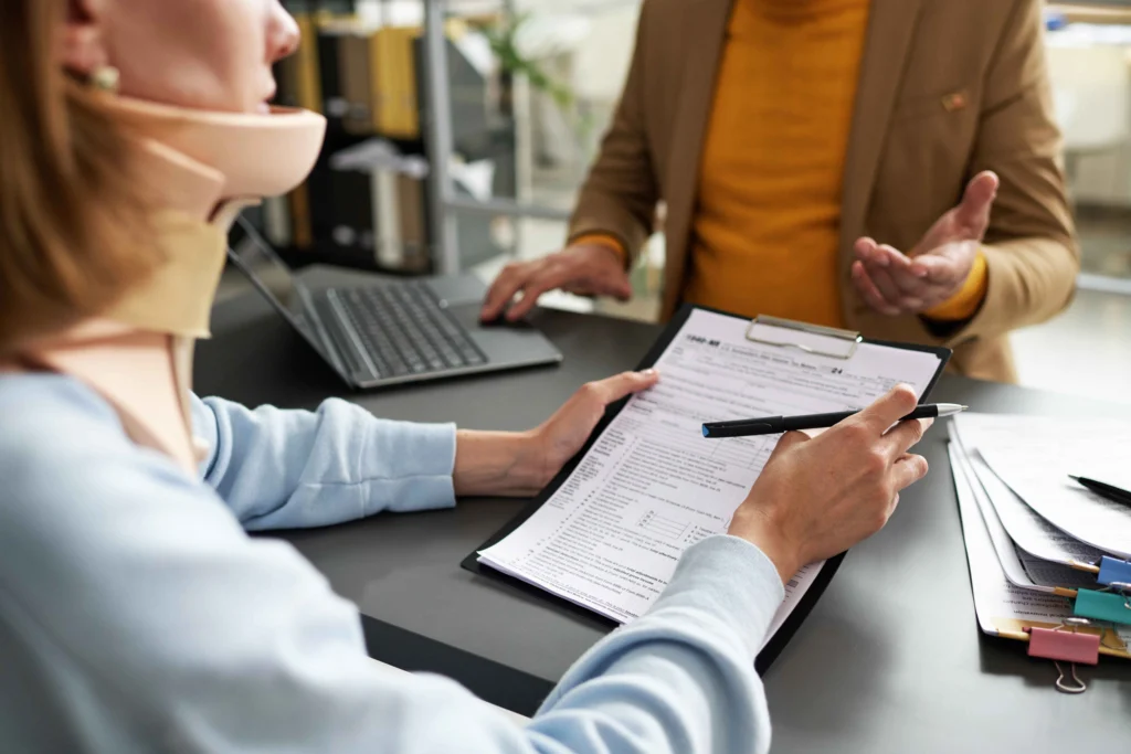 A woman in a neck brace reviews a document and takes notes during a consultation with a person in a brown blazer.
