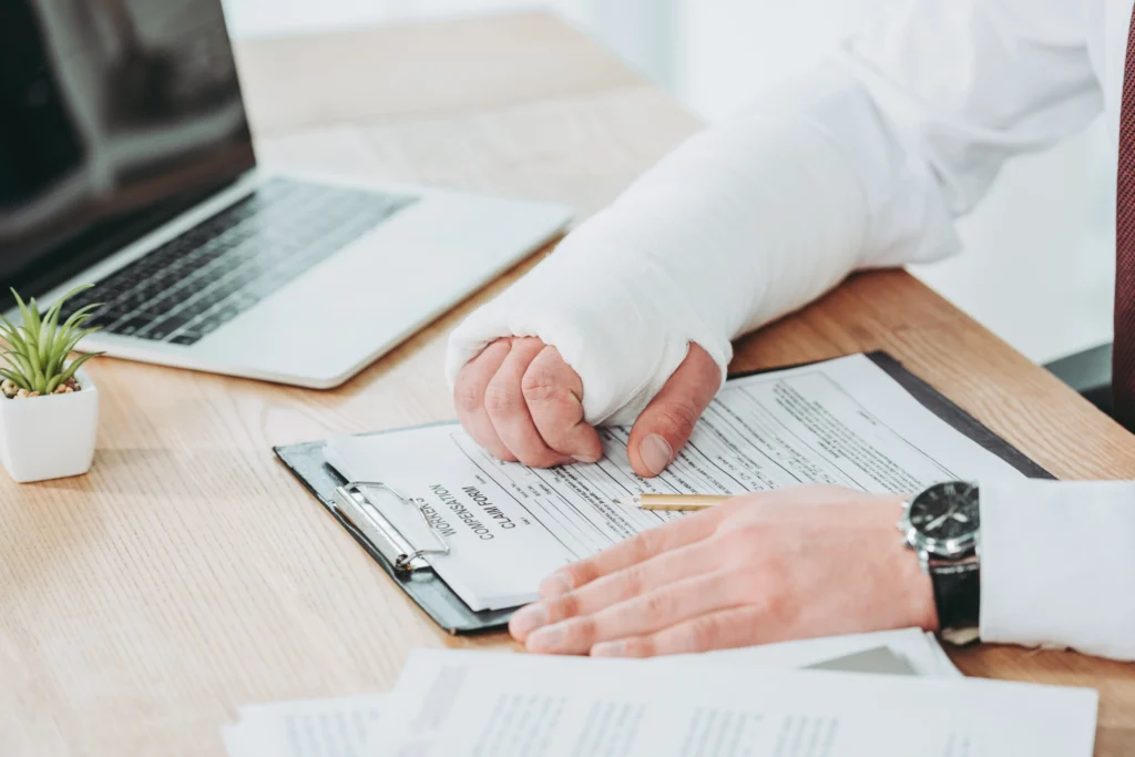 A person with a bandaged hand writes on a clipboard with compensation forms beside a laptop and potted plant on a wooden desk.