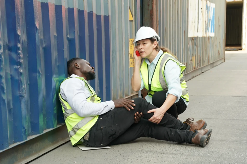 A worker in a safety vest assists an injured colleague sitting against a shipping container, providing immediate care and support.