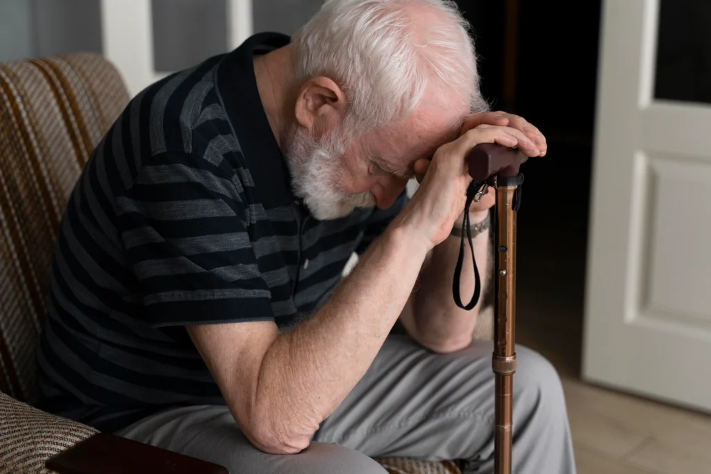 An elderly man sits on a couch, holding a wooden cane, his posture reflecting deep thought or distress.