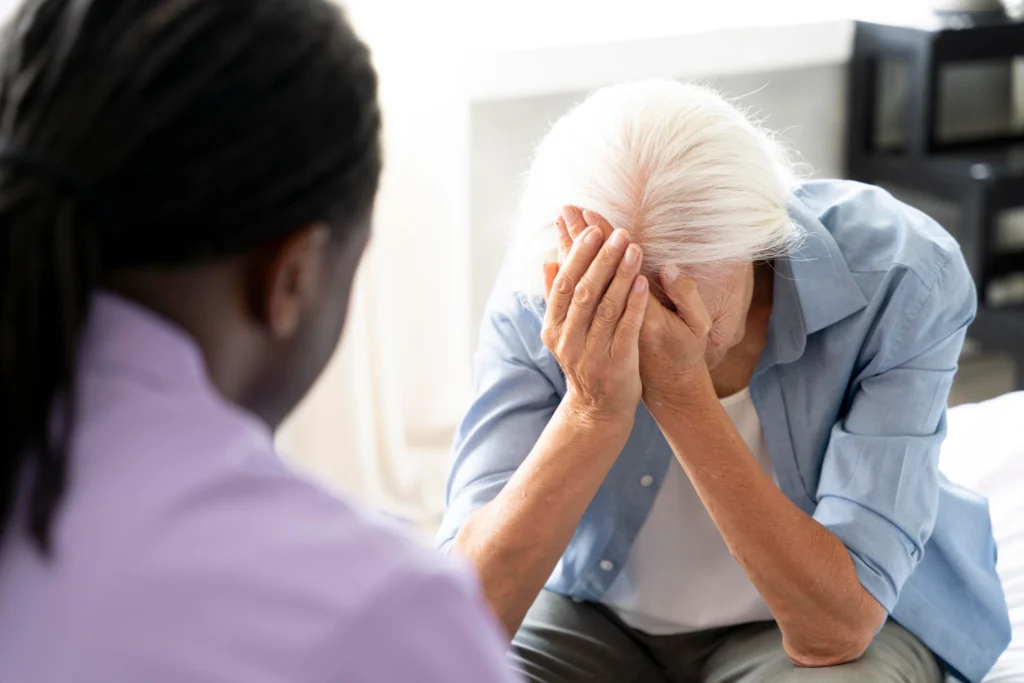 An elderly woman with white hair covers her face in distress while a man in a purple shirt listens empathetically.