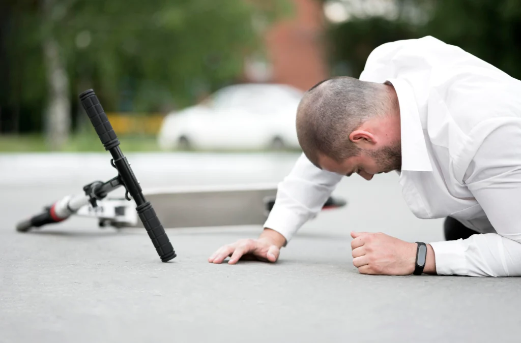 A man in a white shirt lies on the ground next to a fallen scooter, appearing injured or struggling to get up.