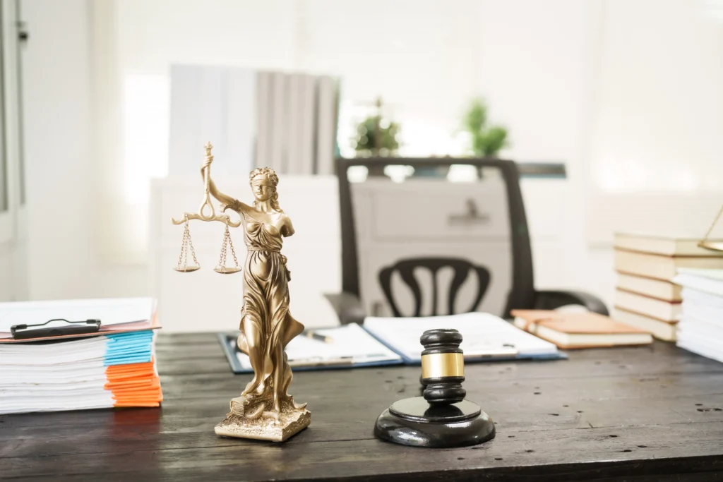 A golden Lady Justice statue scales and a black gavel sit on a dark wooden desk, surrounded by folders and books in a bright office.