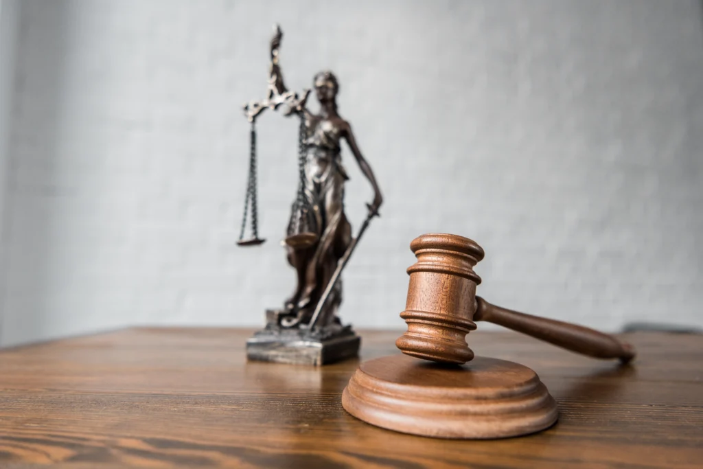 A wooden gavel lies on a table in the foreground, with a statue of Lady Justice holding scales and a sword in the background.