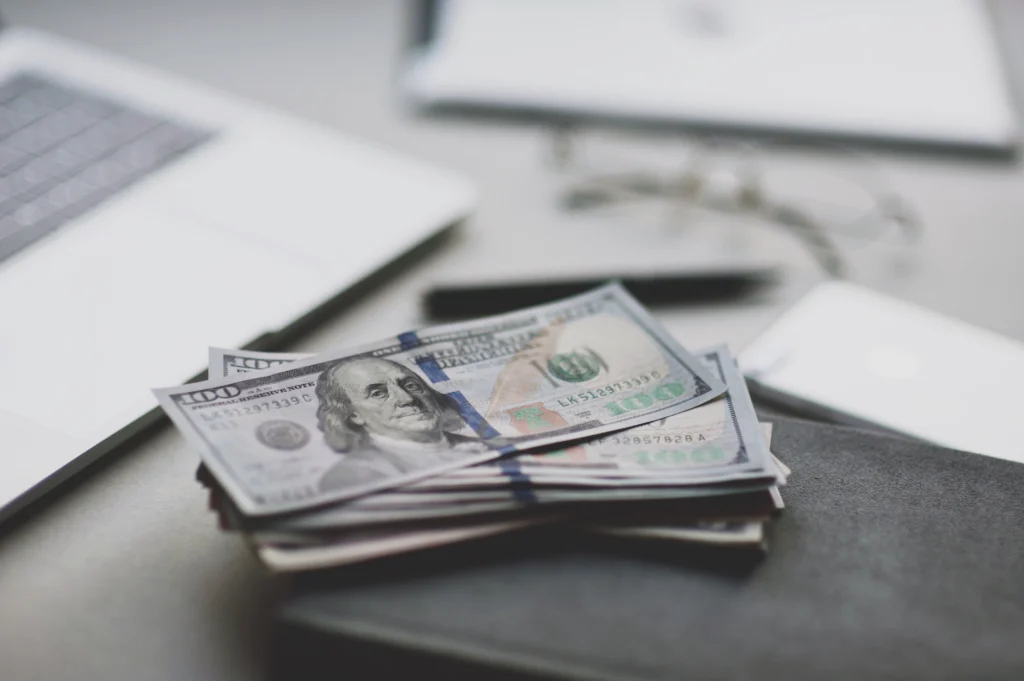 A stack of hundred-dollar bills is placed on a dark notebook, near a laptop and a smartphone. Glasses and a pen are nearby.