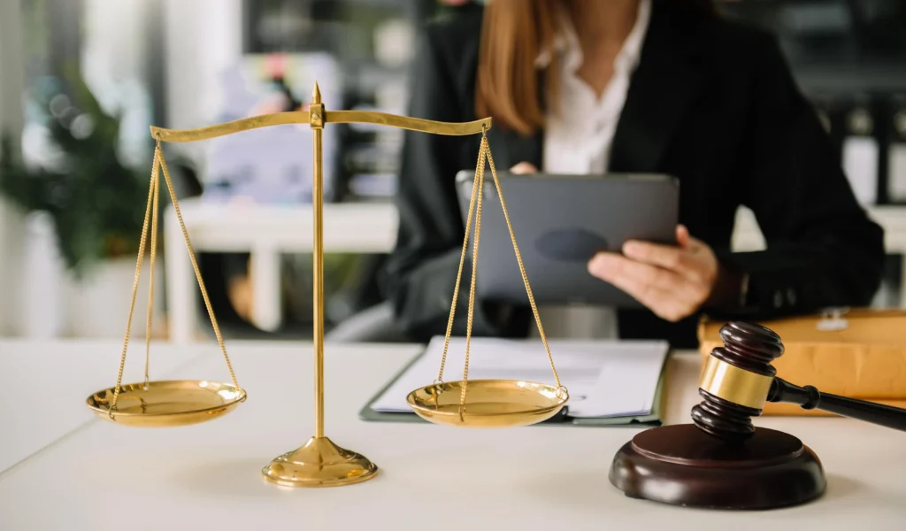 A golden scale of justice stands on a desk next to a gavel, with a professional in a blazer holding a tablet in the background.