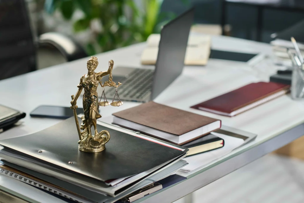 A desk cluttered with files, notebooks, and a laptop, featuring a golden Lady Justice statue holding scales.