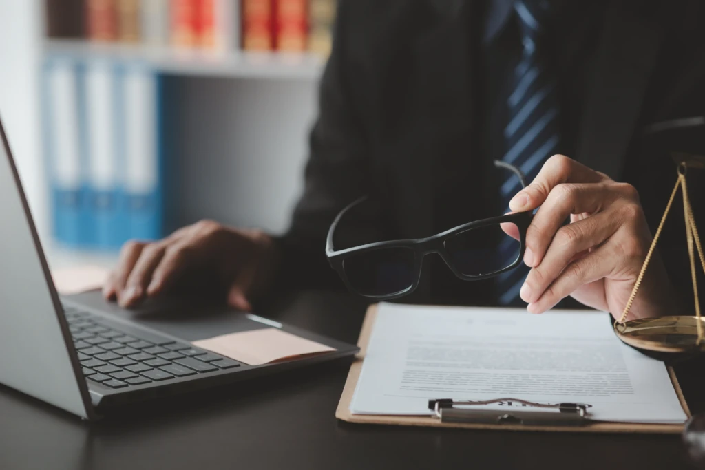 A person in a suit works at a desk with a laptop and clipboard, holding glasses. Scales of justice suggest a legal setting. Books are visible in the background.